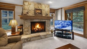 A living room with a stone fireplace, a television on a stand, a ceiling fan, and a view of trees through large windows.