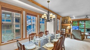 A dining area with a long table set for six, surrounded by wooden chairs. There is a chandelier above the table, and the room has large windows with a view of a stone wall and a staircase.