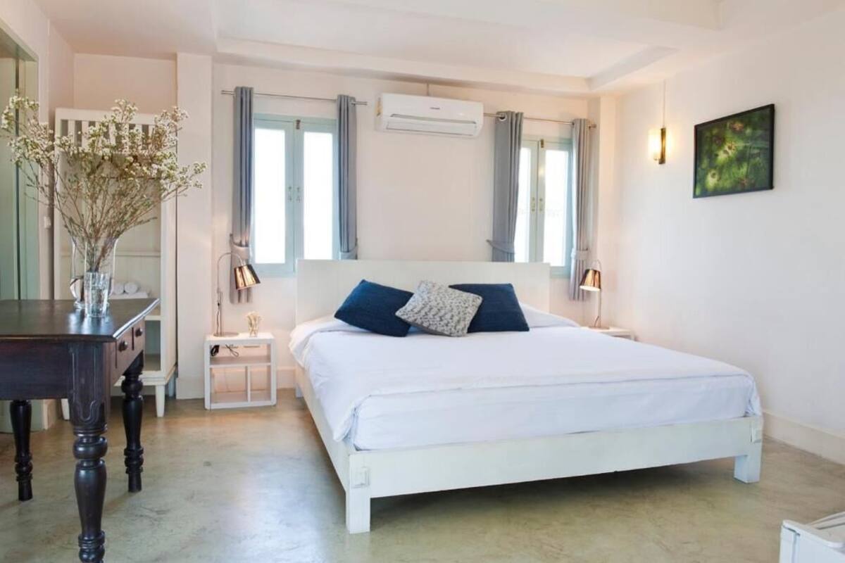 Een slaapkamer met een wit bed, twee kussens, een nachtkastje, een lamp, een airconditioningapparaat en een vaas met bloemen.