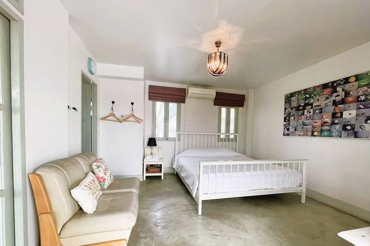 Een slaapkamer met een wit bed, een beige sofa, een klein nachtkastje, een hanglamp en een kunstwerk aan de muur.