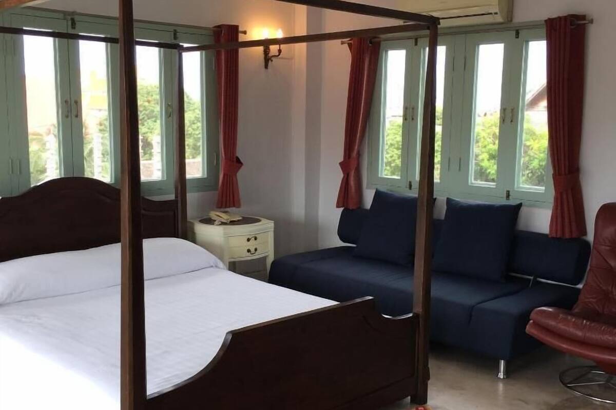 Een hemelbed met een witte matras, een blauwe bank, een rode lederen fauteuil en een kamerkleed met dessin.