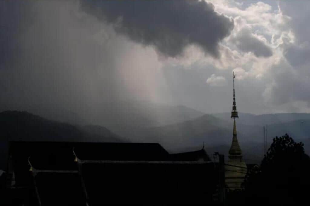 Um edifício tradicional com telhado pontiagudo e torre, com montanhas e céu nublado ao fundo.