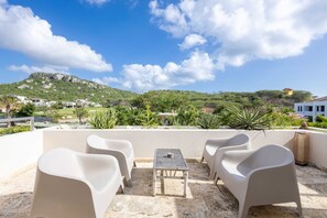 A terrace with white plastic chairs and a table, overlooking a green hillside with buildings. - Seabreeze Retreat Stylish Blue Bay Residence (Sint Michiel)