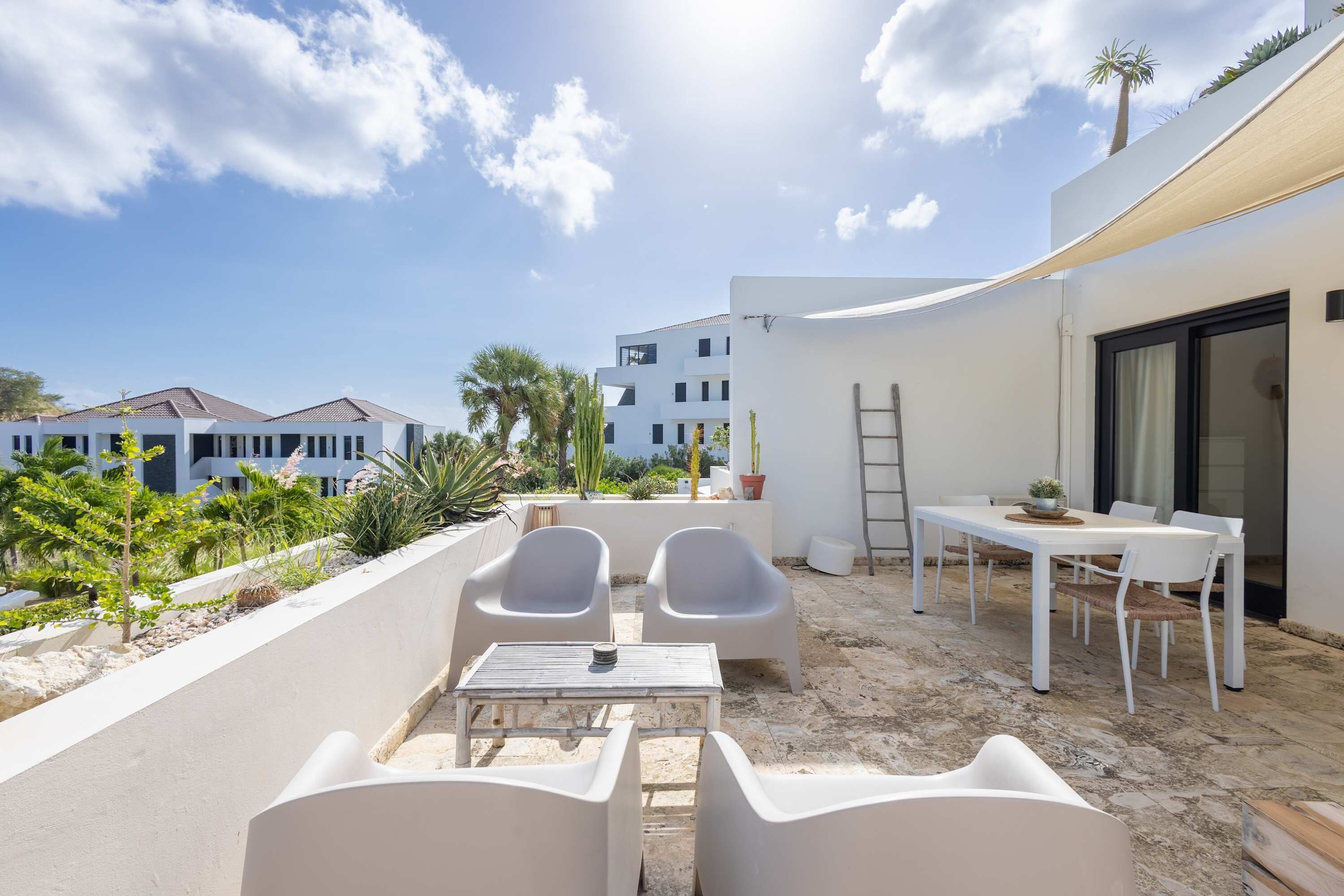 An outdoor patio with white furniture, a table, and chairs, surrounded by a white wall and plants.