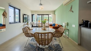 Uma área de refeições com uma mesa redonda, cadeiras de vime, um vaso com flores e vista para o exterior através de amplas janelas.