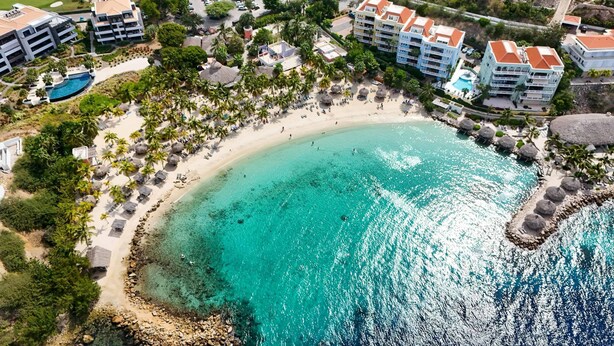Vista aérea de un complejo turístico costero con una playa de arena, aguas turquesas cristalinas y varios edificios con balcones.