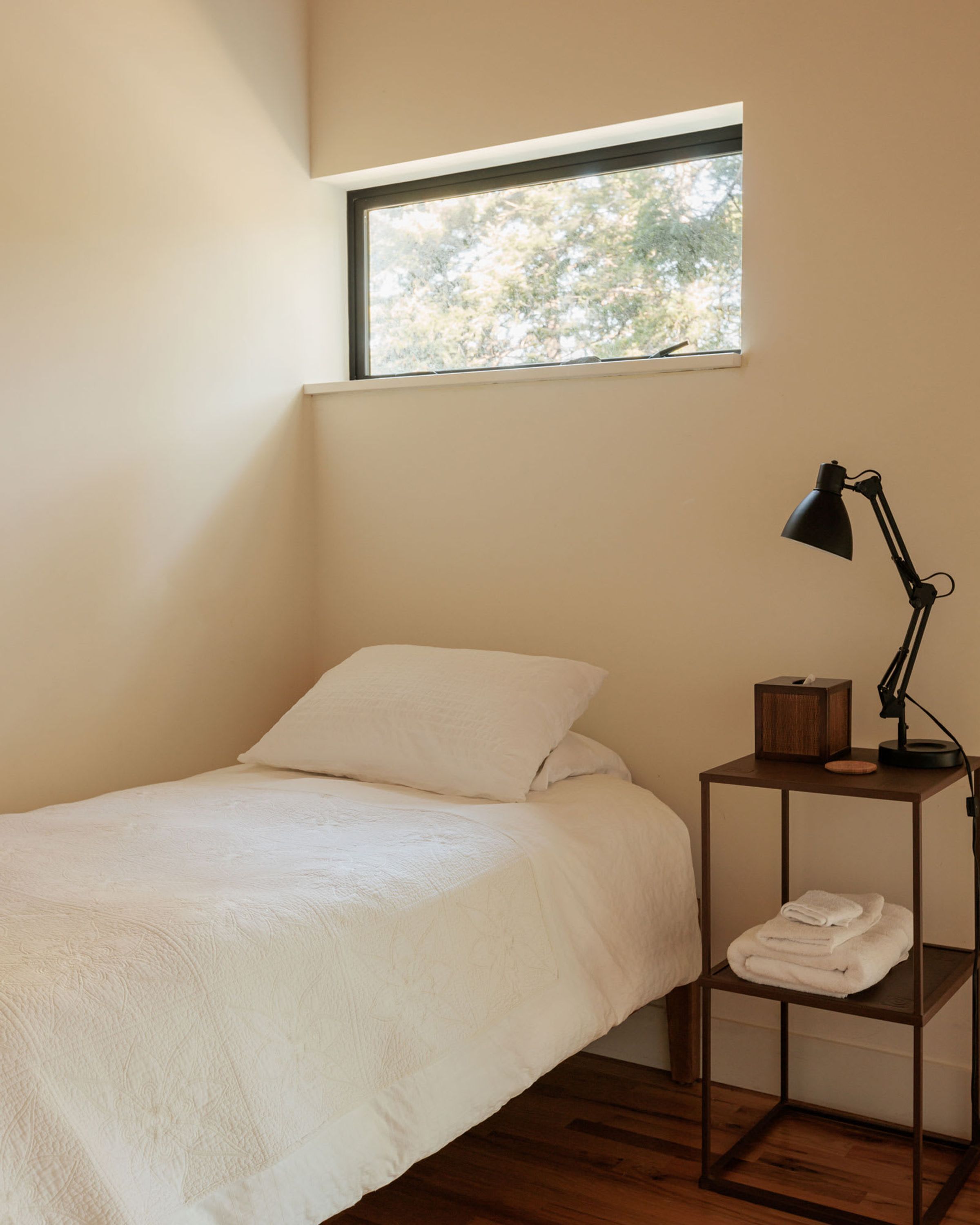 A small, neatly made bed with a white duvet and pillows, a bedside table with a lamp, and a window above the bed.