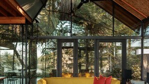 Un interior moderno con piso de madera, un sofá amarillo, sillas verdes y ventanas amplias que dan a unas árboles.
