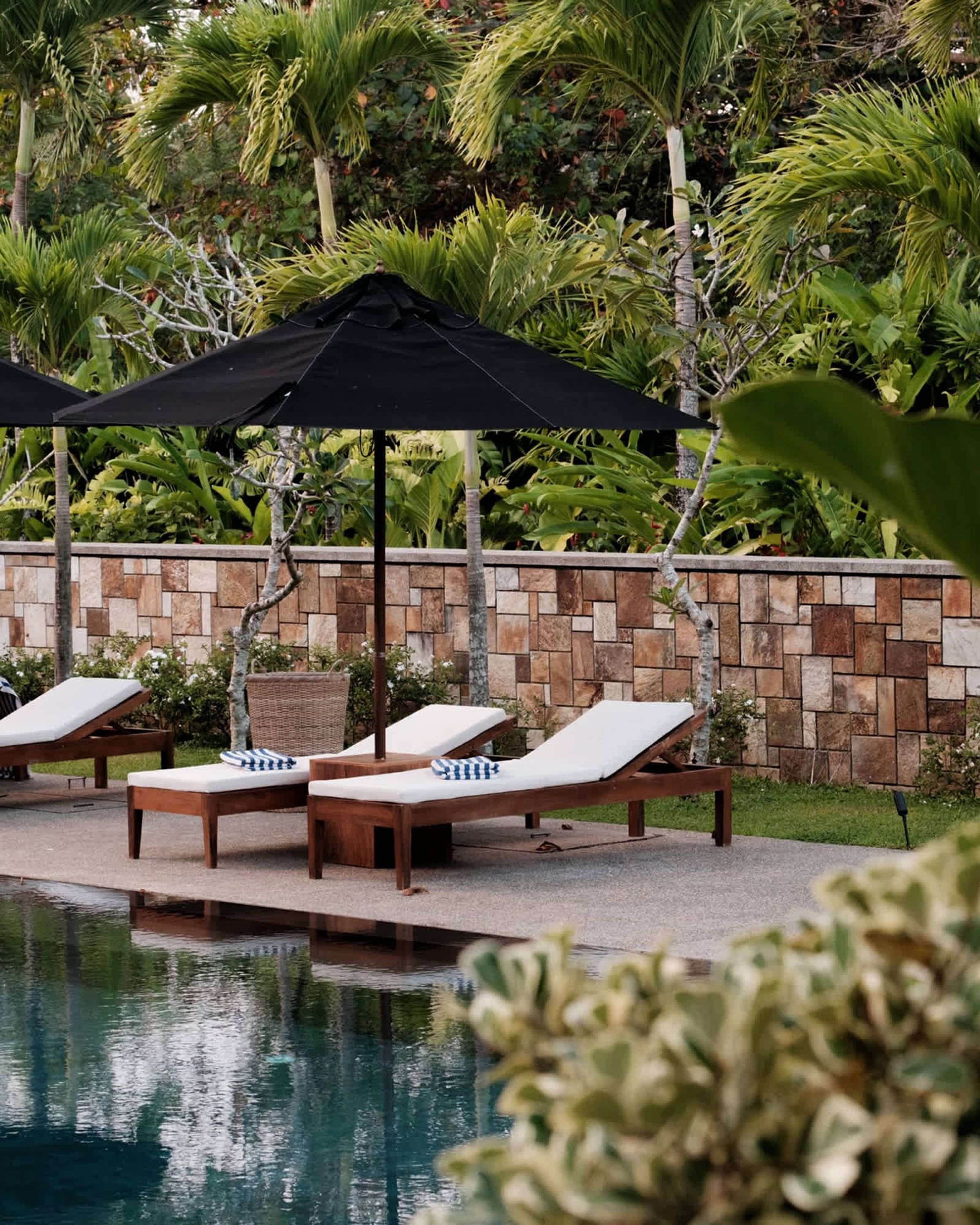Un espace aménagé au bord de la piscine, avec des chaises longues et des parasols, entouré d’un mur de pierre et d’une végétation luxuriante.