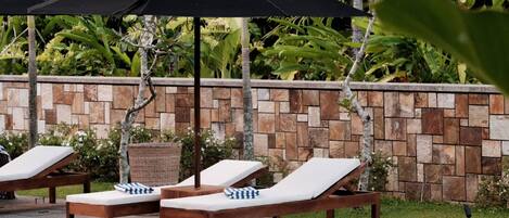 Un espace aménagé au bord de la piscine, avec des chaises longues et des parasols, entouré d’un mur de pierre et d’une végétation luxuriante.