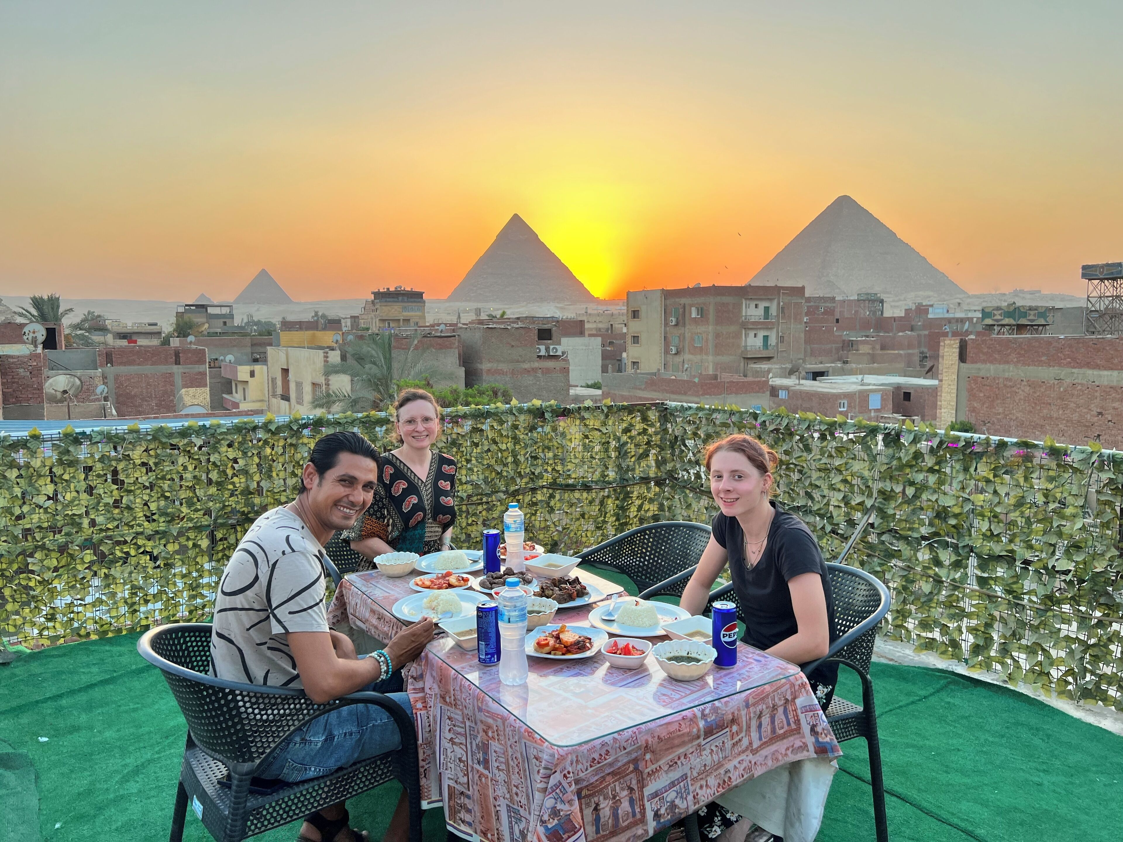Mesa al aire libre con comida y bebidas, con vistas a un paisaje urbano y pirámides al fondo.