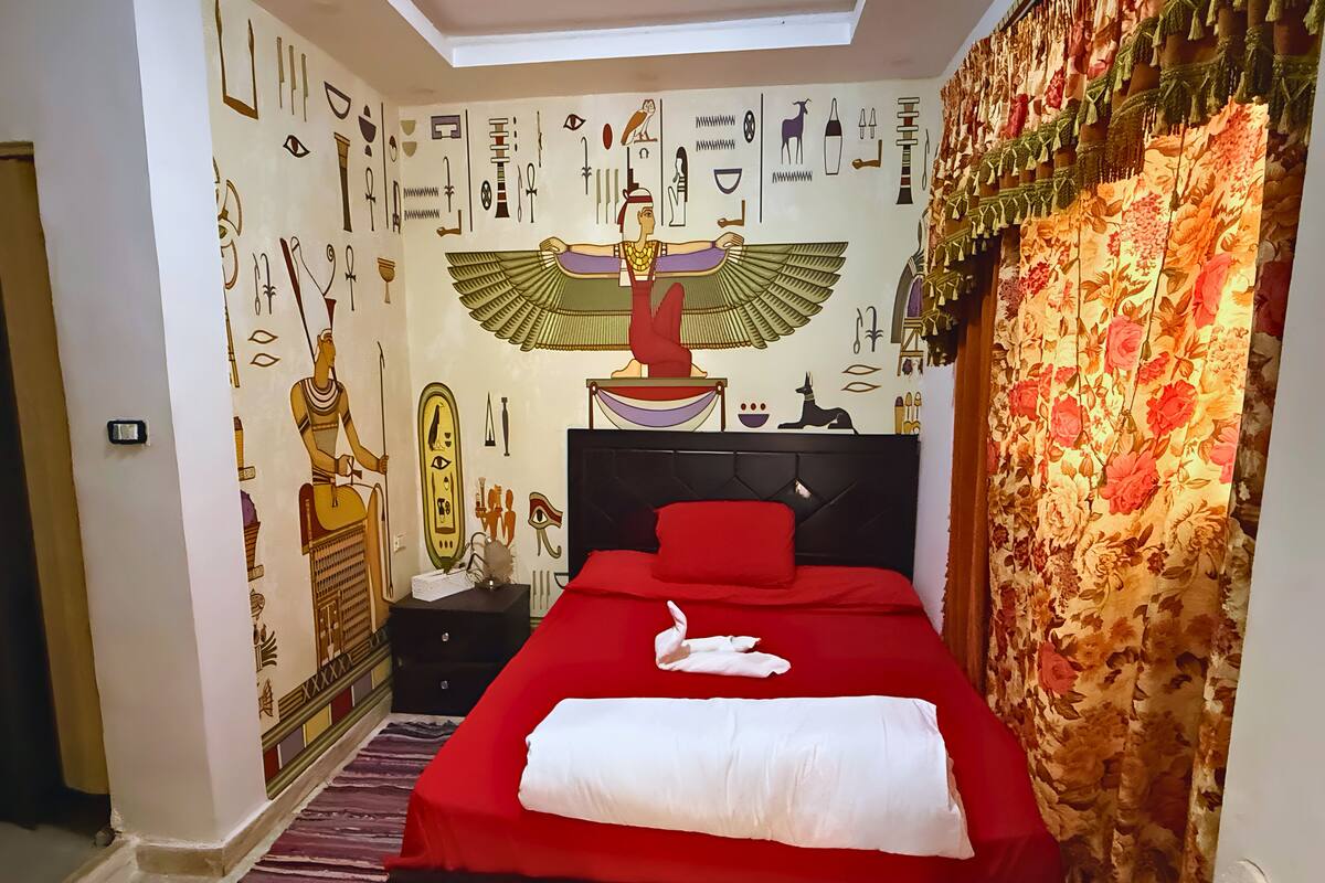 A bedroom with a bed, a nightstand, a wall with Egyptian hieroglyphs, and a window with curtains.