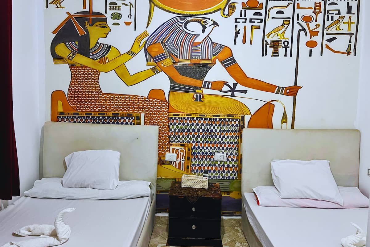 A room with two beds, a wall mural of ancient Egyptian art, and a small table.