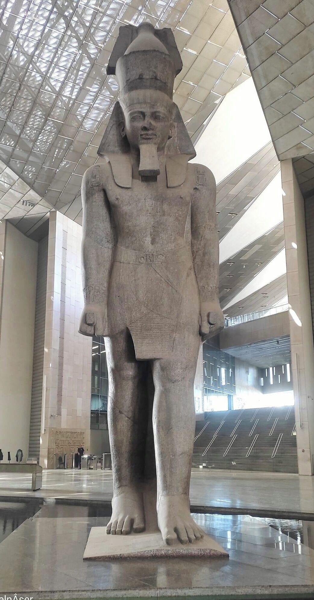 Una gran estatua de piedra de un faraón en un ambiente interior moderno, con techo alto y tragaluces.