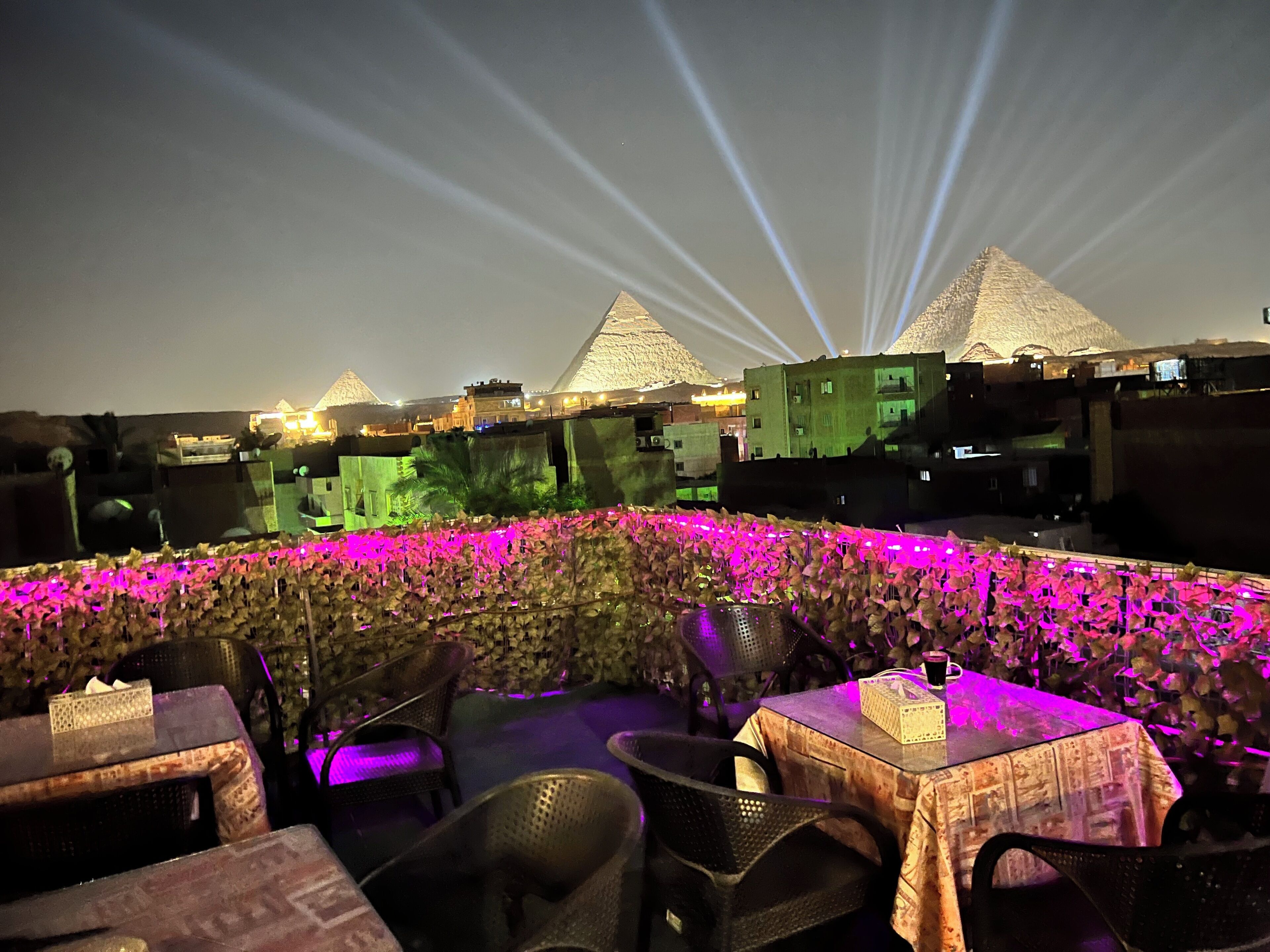 A rooftop terrace with tables and chairs, a decorative wall, and a view of pyramids in the background.