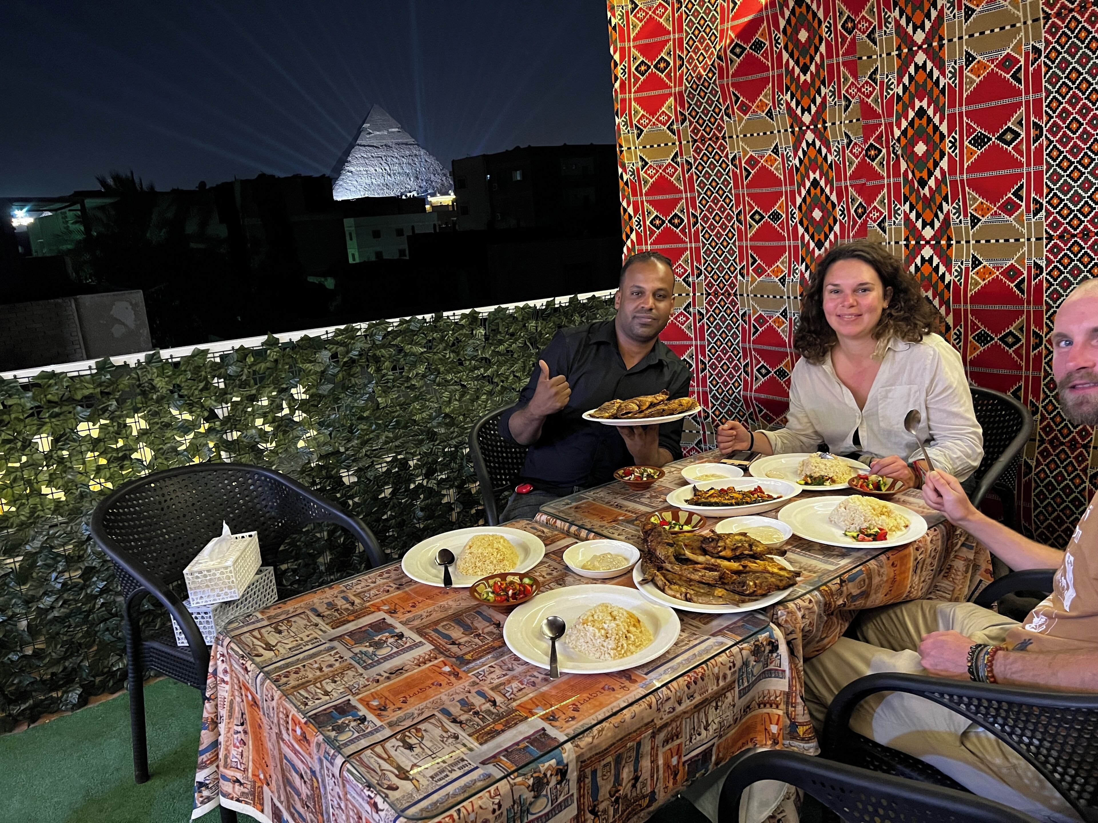 Une table à manger, sur laquelle sont disposés de nombreux plats, entourée de chaises, le tout se détachant sur un fond de tissu à motifs et surmonté d’une structure lumineuse en forme de pyramide.