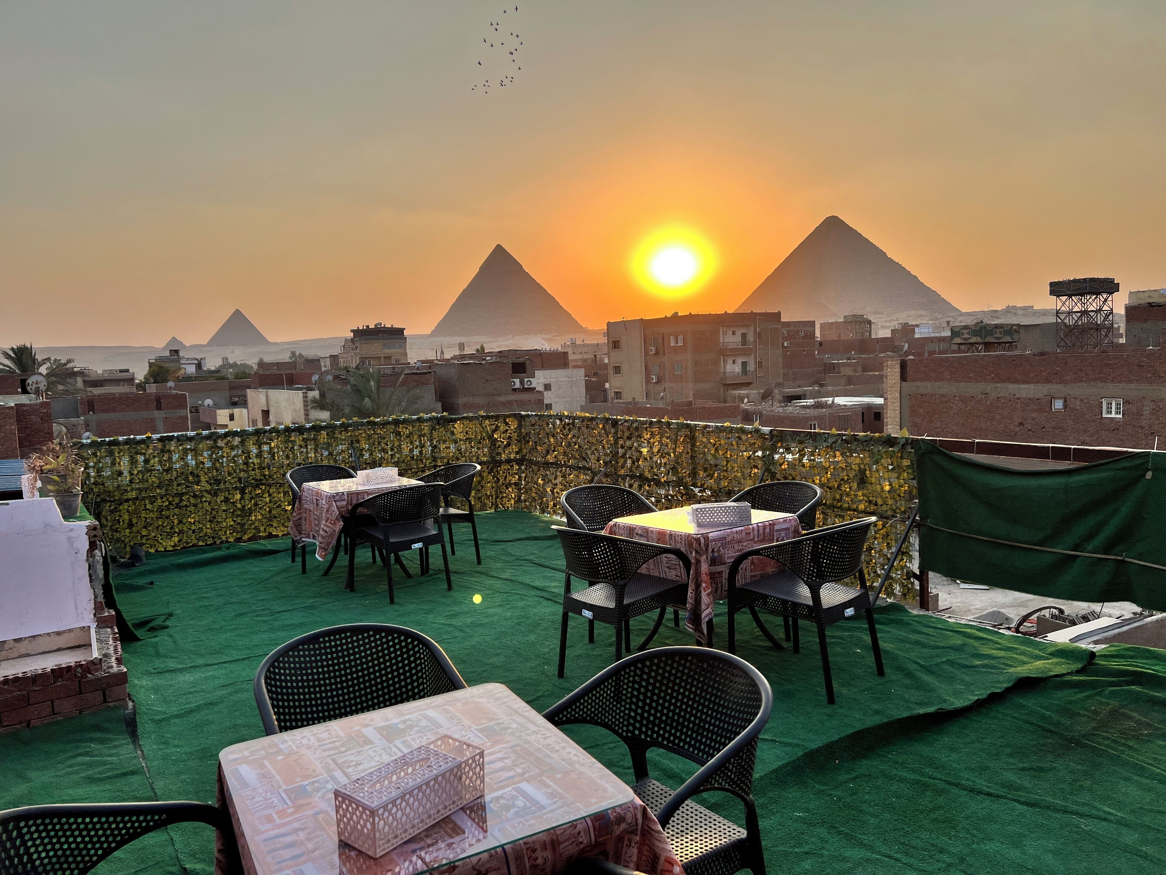 Terraza en la azotea con mesas y sillas, con vistas a un paisaje urbano y pirámides al atardecer.