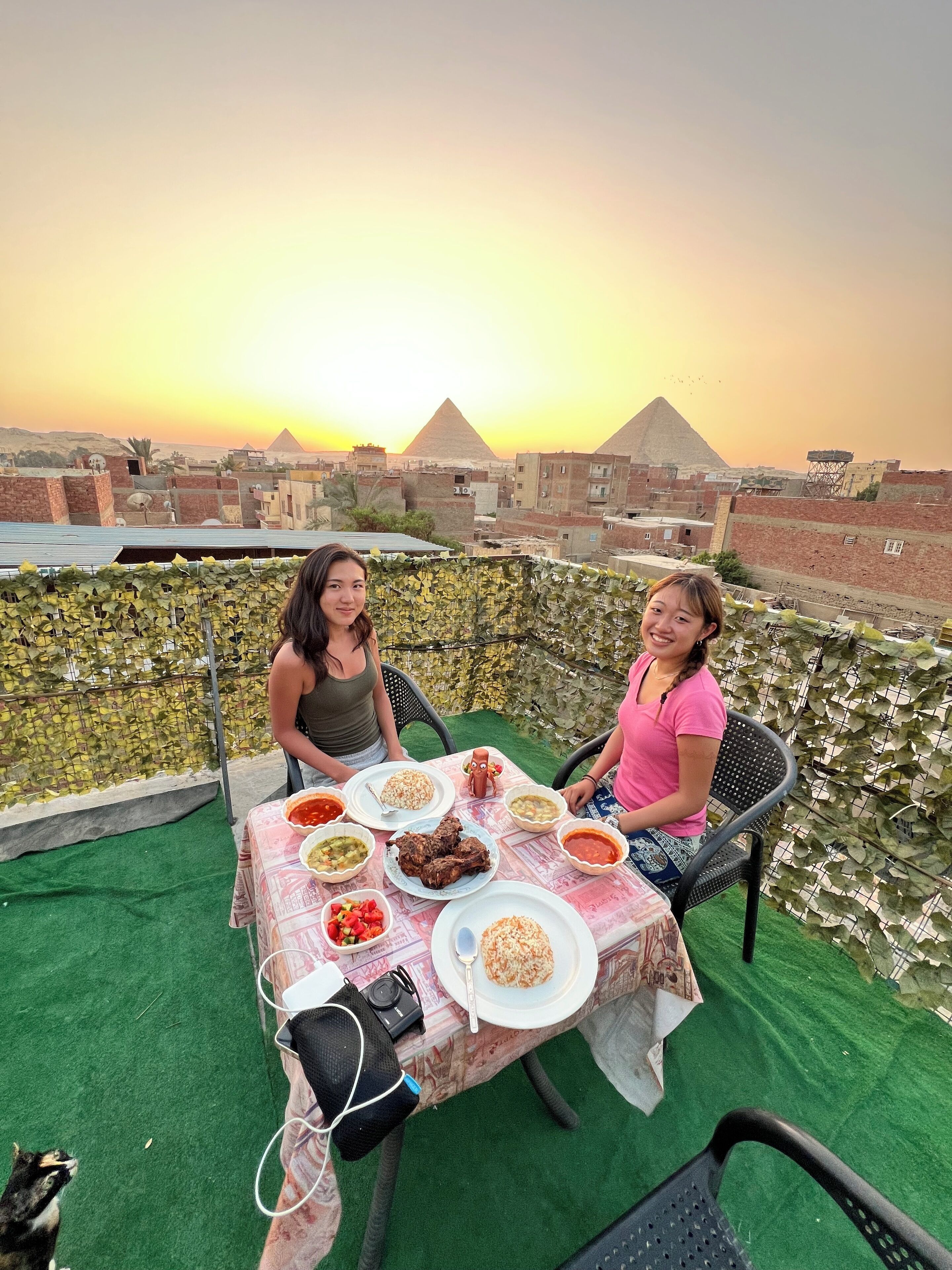 Zwei Personen essen an einem Tisch mit verschiedenen Gerichten, mit Blick auf eine Stadtlandschaft und Pyramiden im Hintergrund.