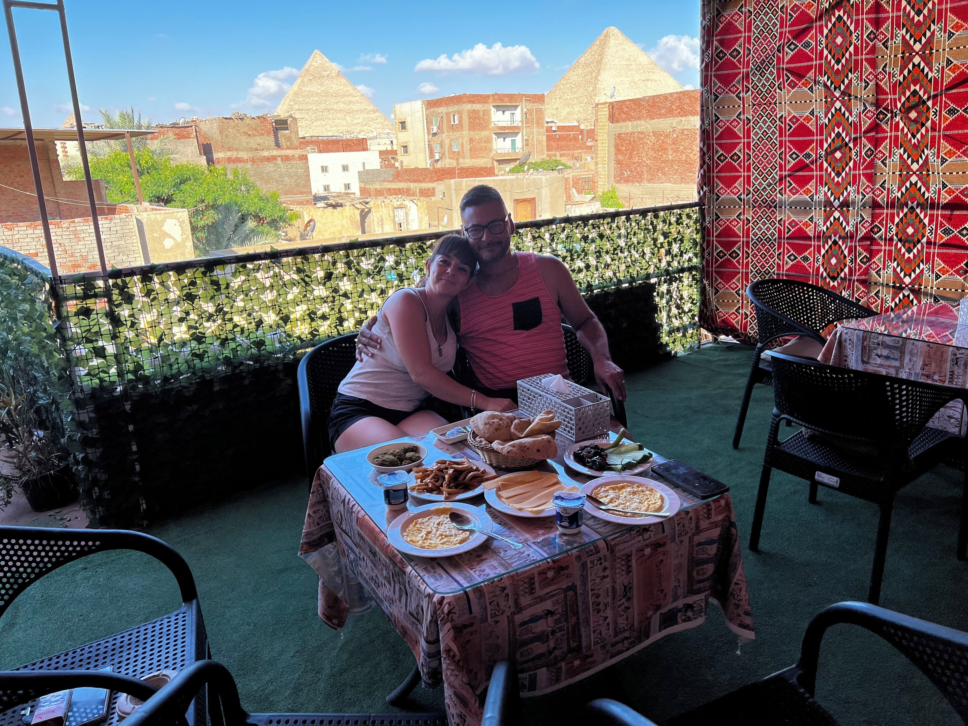 Ein Paar speist auf einem Balkon mit Blick auf Pyramiden, einem gemusterten Tischlein und einem Brotkorb.