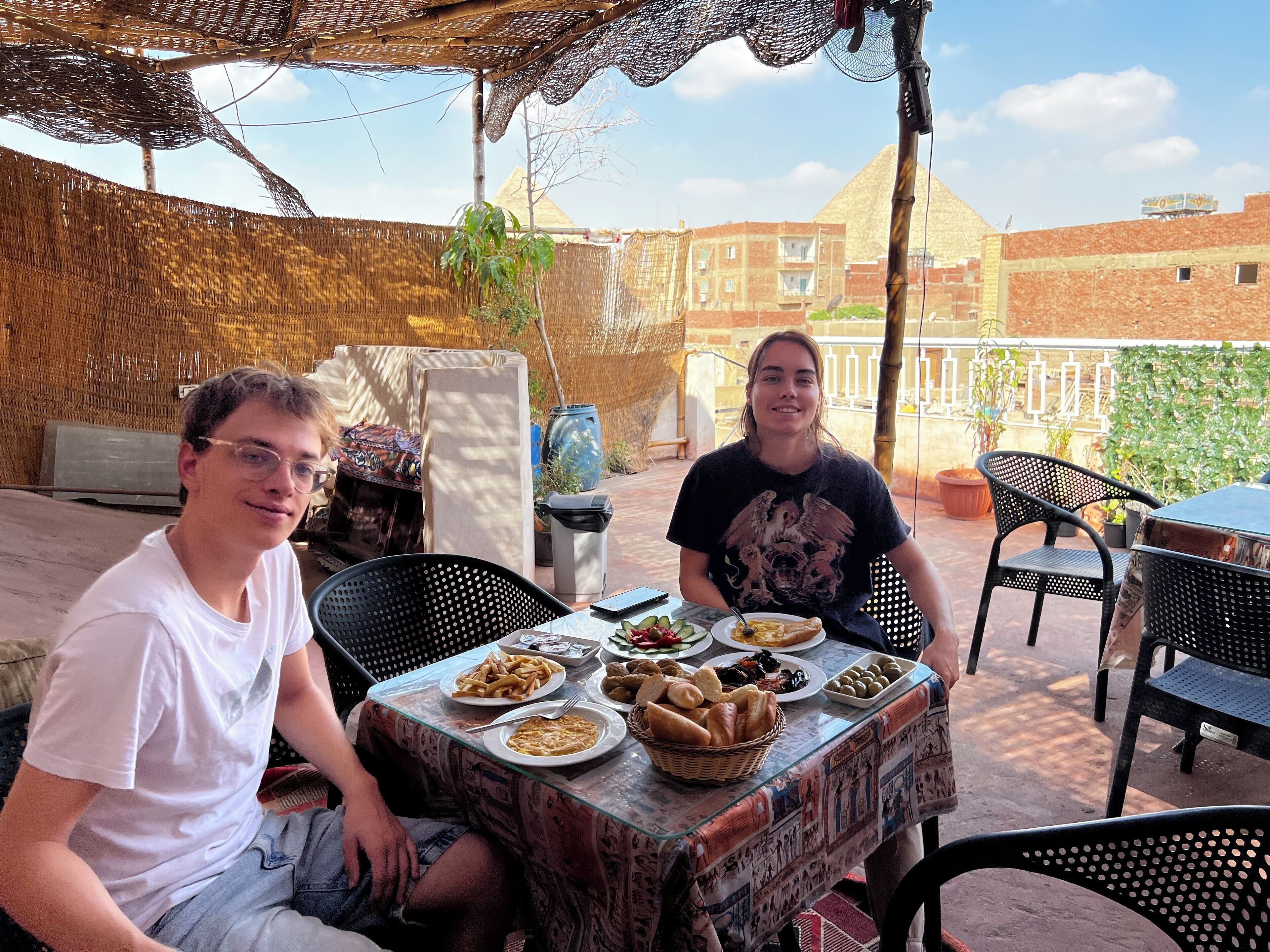 Two people seated at an outdoor table with various dishes, under a shaded area with a woven structure.