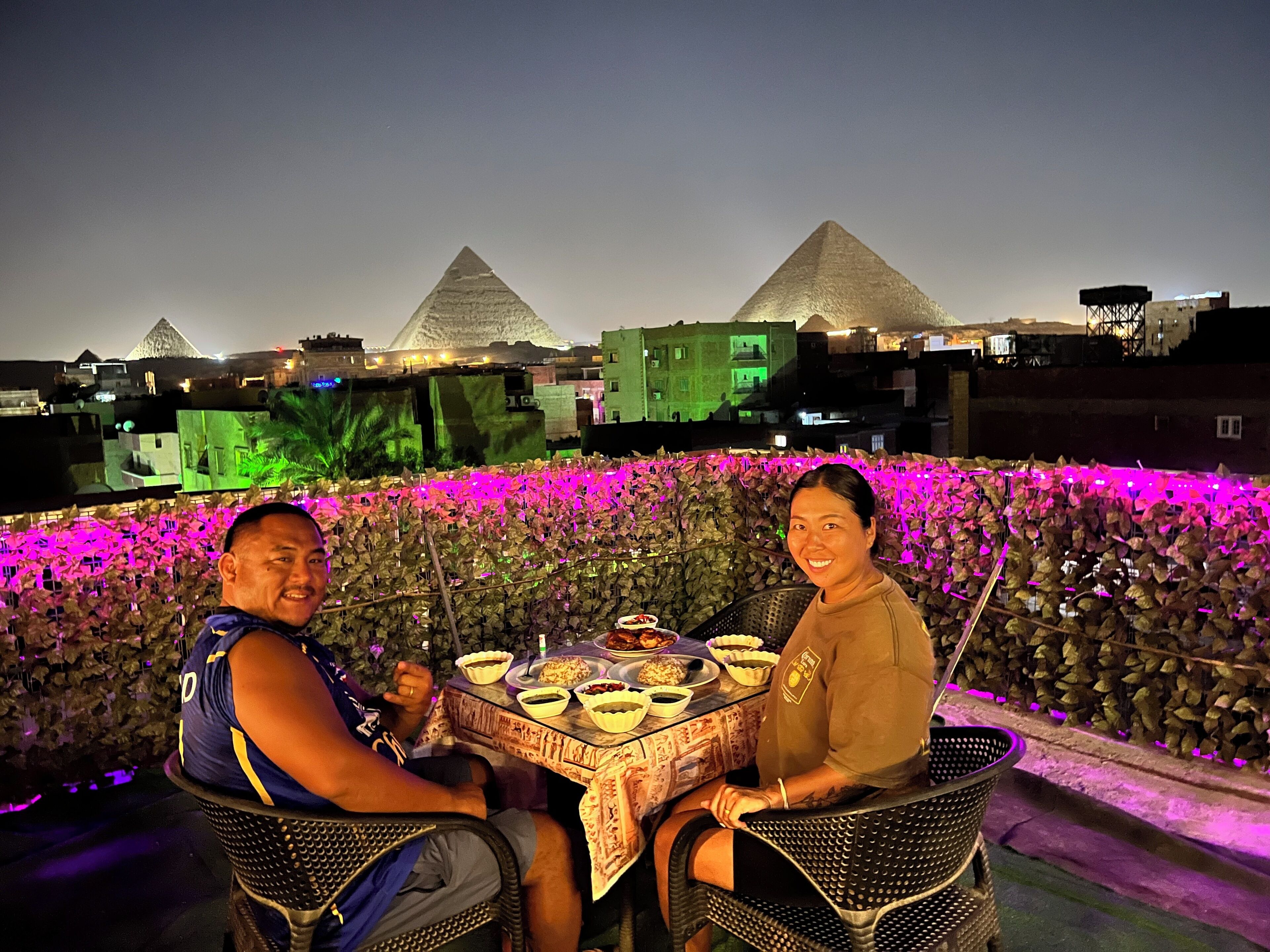 A dining table with food and drinks, surrounded by chairs, with pyramids in the background.