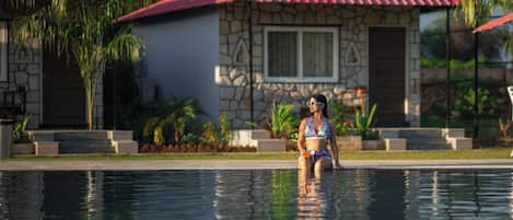 Una mujer sentada junto a una piscina, con edificios de piedra y tejados rojos al fondo.