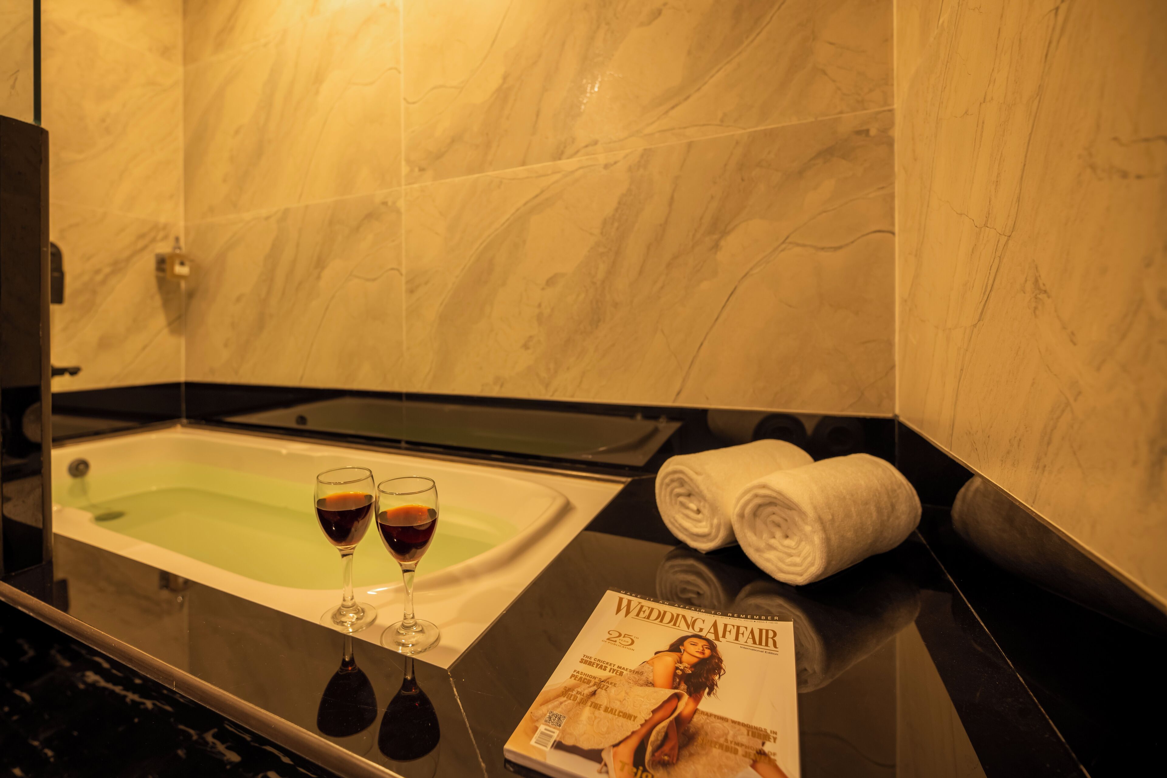 Une salle de bain avec une baignoire, deux verres de vin rouge et un magazine posé sur le comptoir.