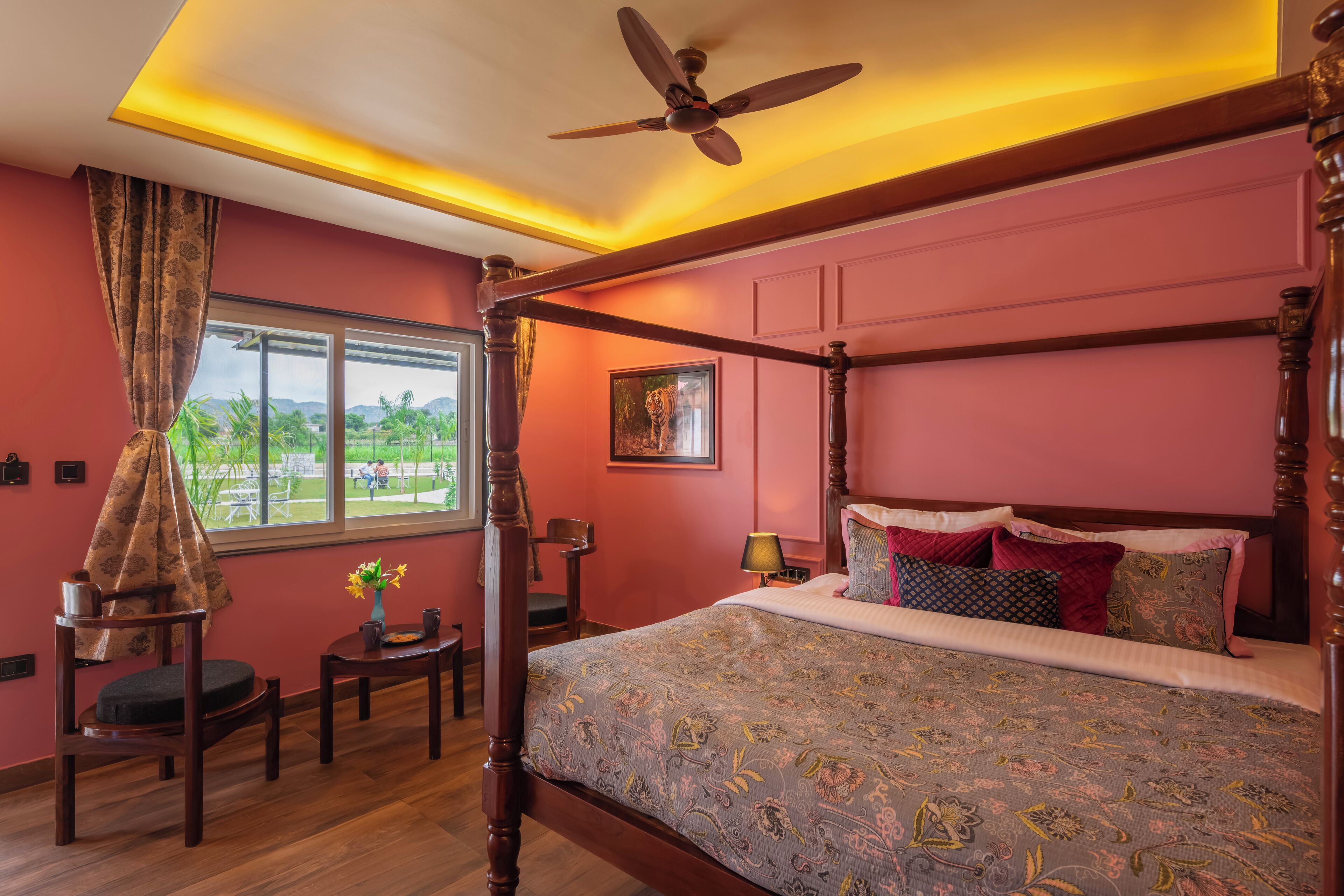 A four-poster bed with a patterned bedspread, a ceiling fan, a window with a view of greenery, and a small table with a vase of flowers.