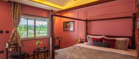 A four-poster bed with a patterned bedspread, a ceiling fan, a window with a view of greenery, and a small table with a vase of flowers.