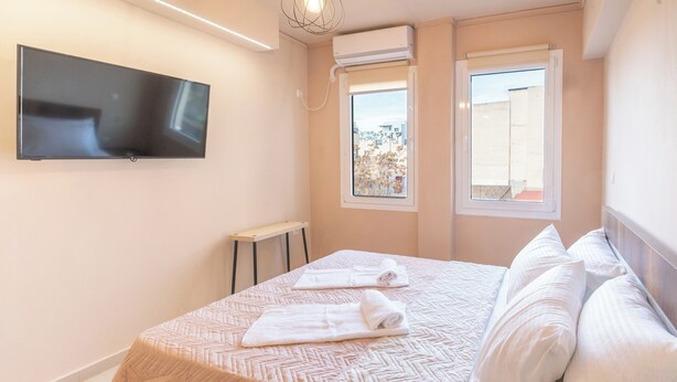 A bedroom with a bed, a flat-screen TV mounted on the wall, two windows, and an air conditioning unit. - Monastiraki metro st studio for 2 persons by MPS (Athina)