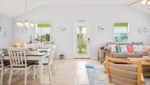 Un vaste espace de vie comprenant une table à manger, des chaises, un canapé et une table basse. Une porte donne accès à un balcon, et la pièce présente un design moderne avec un ventilateur de plafond et des œuvres d’art murales.