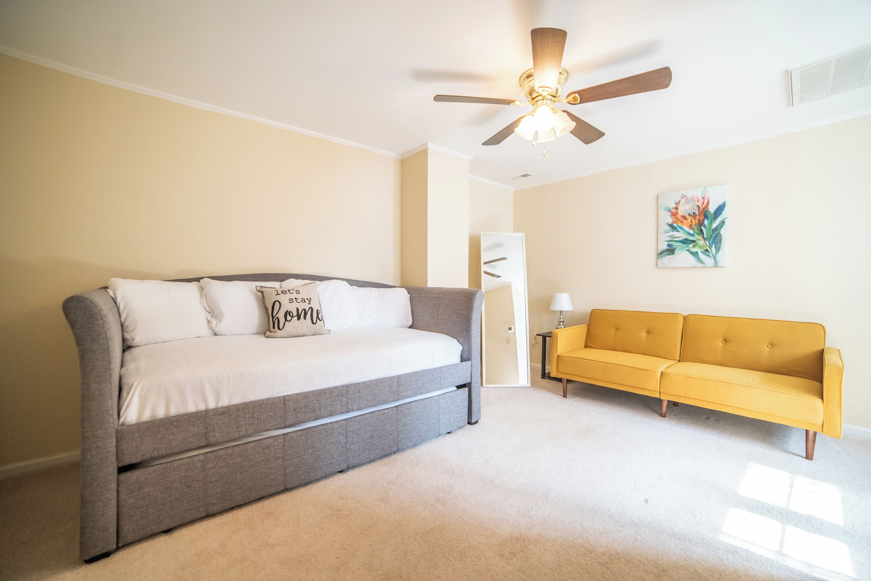 A bedroom with a gray sofa, a yellow sofa, a ceiling fan, and a painting on the wall.