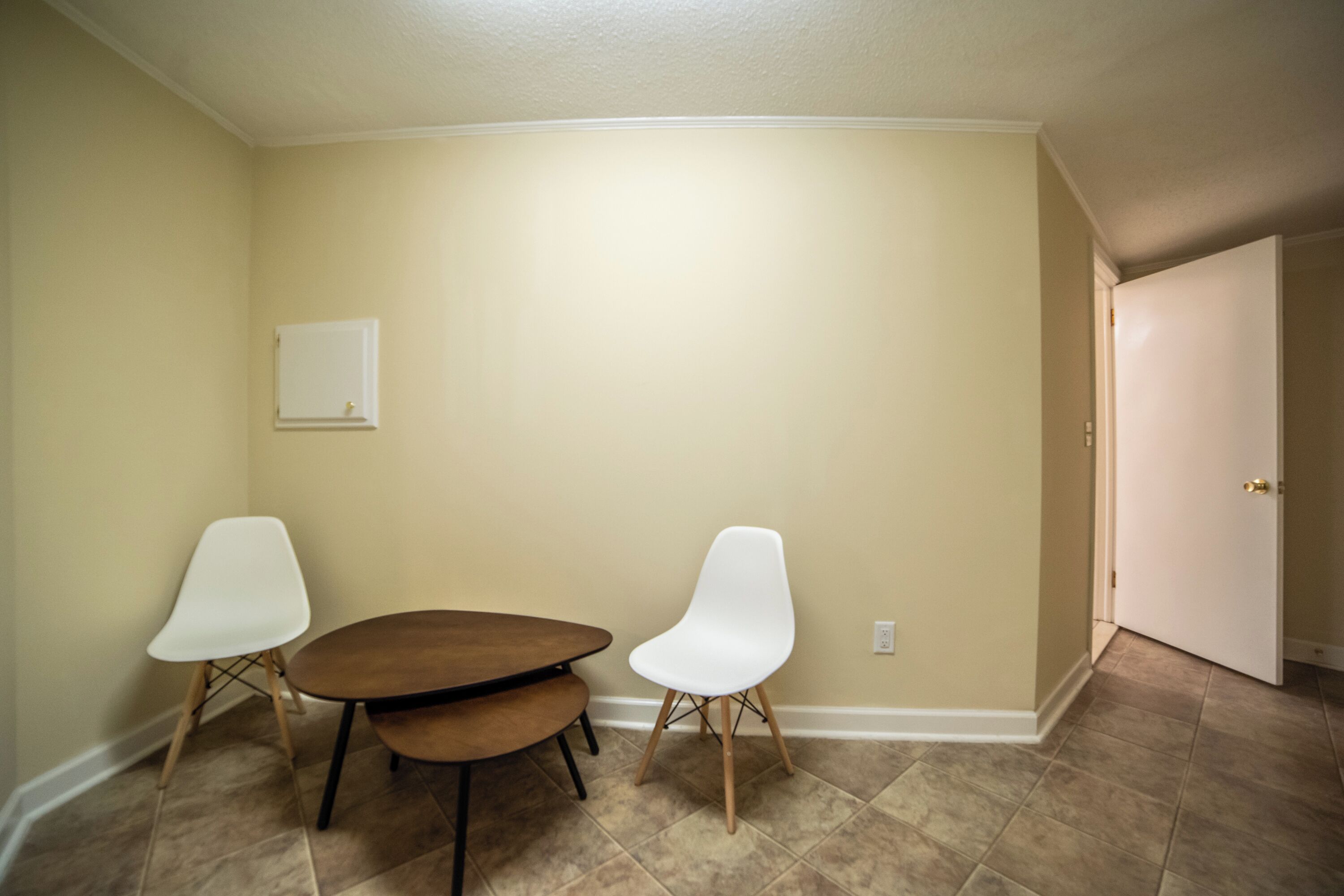Une pièce aux murs de couleur beige, avec une table en bois, deux chaises blanches et une porte blanche.