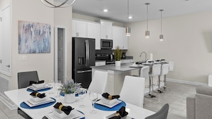 A modern kitchen with white cabinets, a black refrigerator, and a microwave. There is a dining area with a white table set for four, blue placemats, and white plates. - Modern 4BR Pool Townhome | Pet-Friendly 361 (Kissimmee)