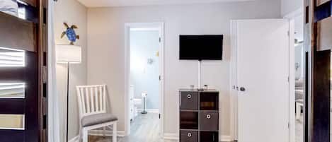 Un pasillo con una puerta blanca, un televisor de pantalla plana montado en la pared, una silla blanca y una lámpara de pie.