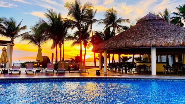 A poolside area with lounge chairs and a thatched-roof pavilion, overlooking a sunset over the ocean. - Beach and luxury with ideal location (Mazatlán)