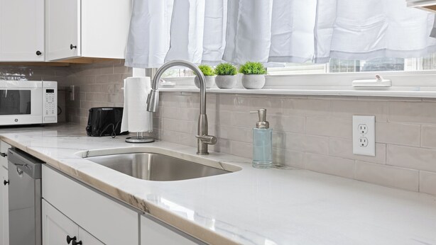 Una cocina moderna con armarios blancos, un lavamanos de acero inoxidable, un microondas y una ventana con cortinas blancas.