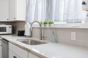 A modern kitchen with white cabinets, a stainless steel sink, a microwave, and a window with white curtains. - Trendy at Hoyt Two Homes (Chattanooga)