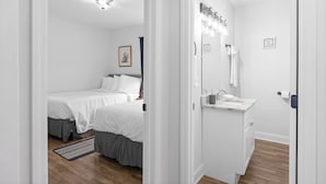 Un dormitorio con dos camas, un baño con lavamanos y espejo, y un pasillo.