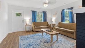 Sala de estar con dos sofás, una mesa de centro y un ventilador de techo. Hay dos ventanas con cortinas azules, una puerta y una chimenea.
