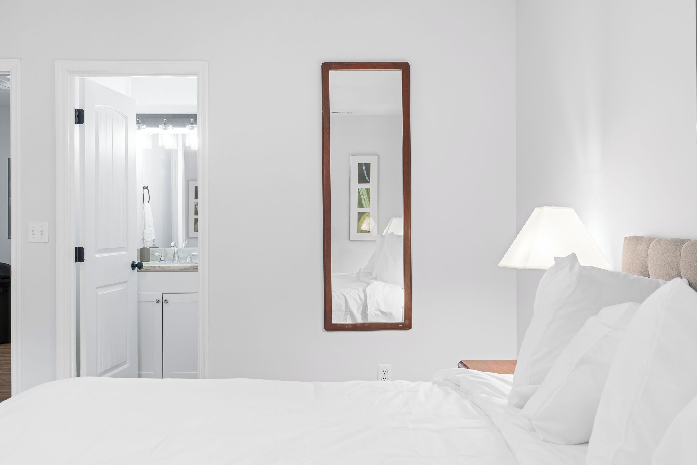 Habitación de hotel con cama, mesita de noche con lámpara, espejo en la pared y puerta que da a un baño.
