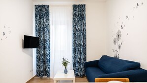 Un salon moderne avec un canapé bleu, un téléviseur à écran plat fixé au mur, une petite table blanche ornée d’un vase de fleurs et un coin repas avec une table dressée pour deux personnes.