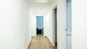 Un couloir étroit doté d’un plancher en bois, de murs blancs et d’une porte donnant sur une pièce où se trouve un canapé bleu.