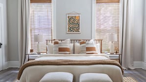 Quarto com uma cama grande, duas mesas de cabeceira, uma obra de arte na parede e uma janela com persianas.