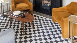 Uma sala de estar com lareira preta, dois poltronas cor mostarda, uma mesa de centro redonda e um tapete com padrão preto e branco.