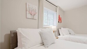 A hotel room with two single beds, a window with blinds, a wall-mounted artwork, and a nightstand with a lamp. - Columbia Duet Unit B (West Columbia)