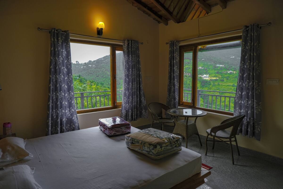 Quarto com cama, mesa, cadeiras, varanda com vista para o verde e janela com cortinas.