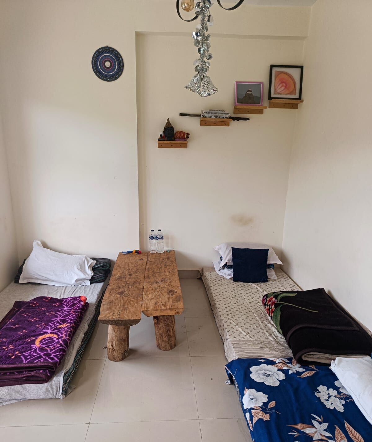 A small, cozy room with two single beds, a wooden table, and a shelf with decorative items.