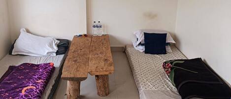 A small, cozy room with two single beds, a wooden table, and a shelf with decorative items.