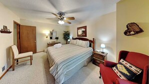 A hotel room with a bed, a chair, a desk, a nightstand, a lamp, a ceiling fan, a mirror, a door, and a wall-mounted shelf. - Pinon Park C11 | Cozy, Fireplace, Great Location (Ruidoso)